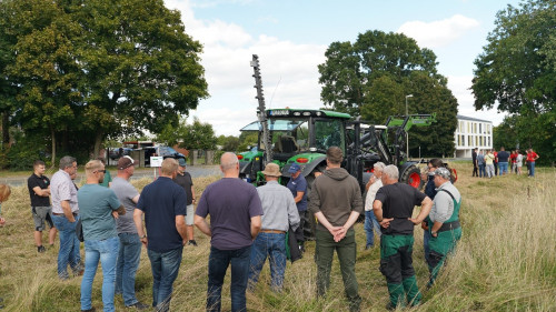 Menschen stehen auf einem Feldrandstreifen und sehen einem Traktor mit Mähwerk an.