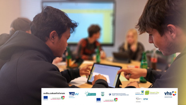 Zwei junge Menschen am Tablet bei kollaborativer Gruppenarbeit, im Hintergrund weitere Personen und ein Digiboard